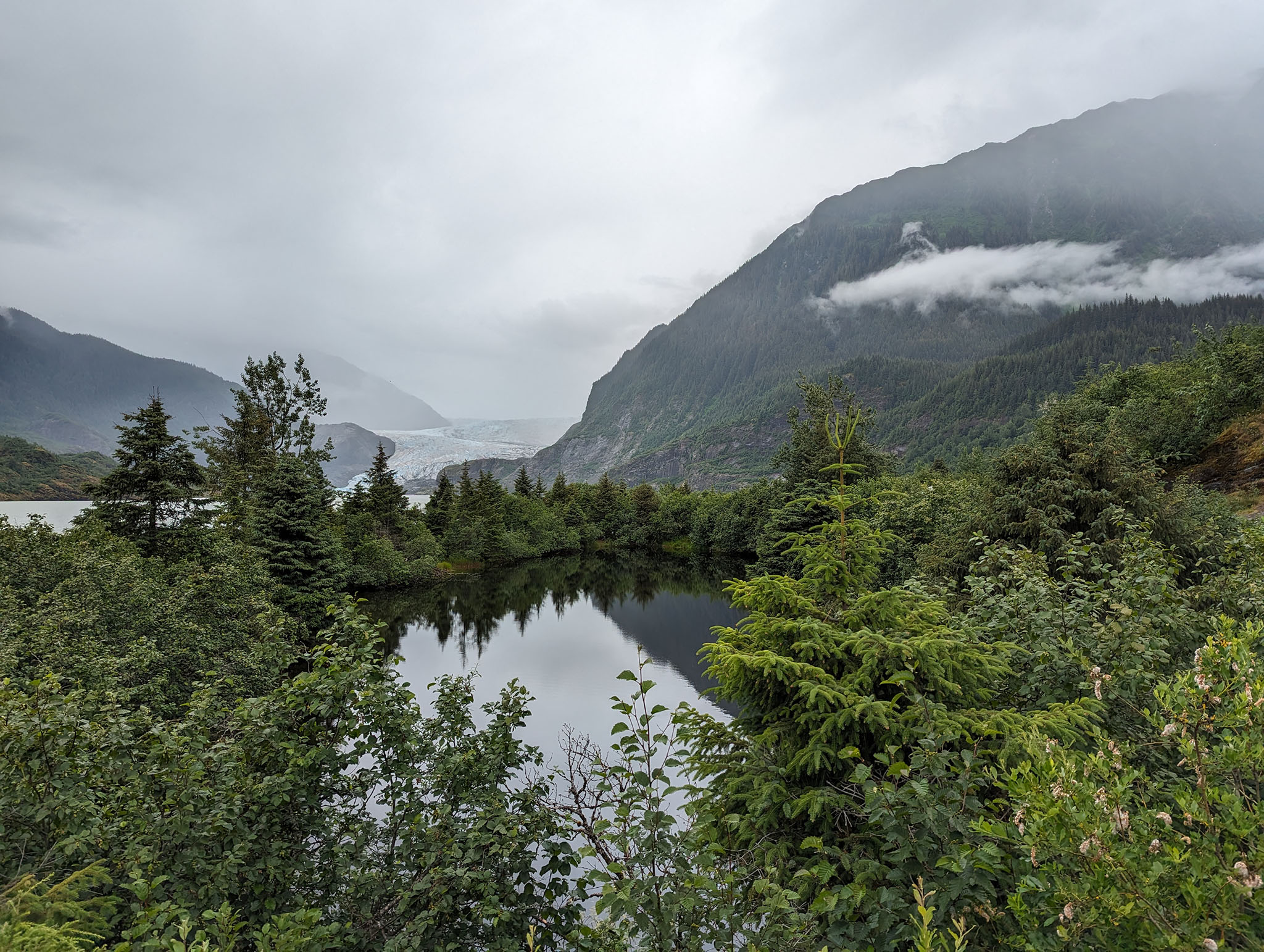 Low cloud over a mountainous landscape with a glacier visible between ridges and a foreground of trees and a flat lake reflecting the scene