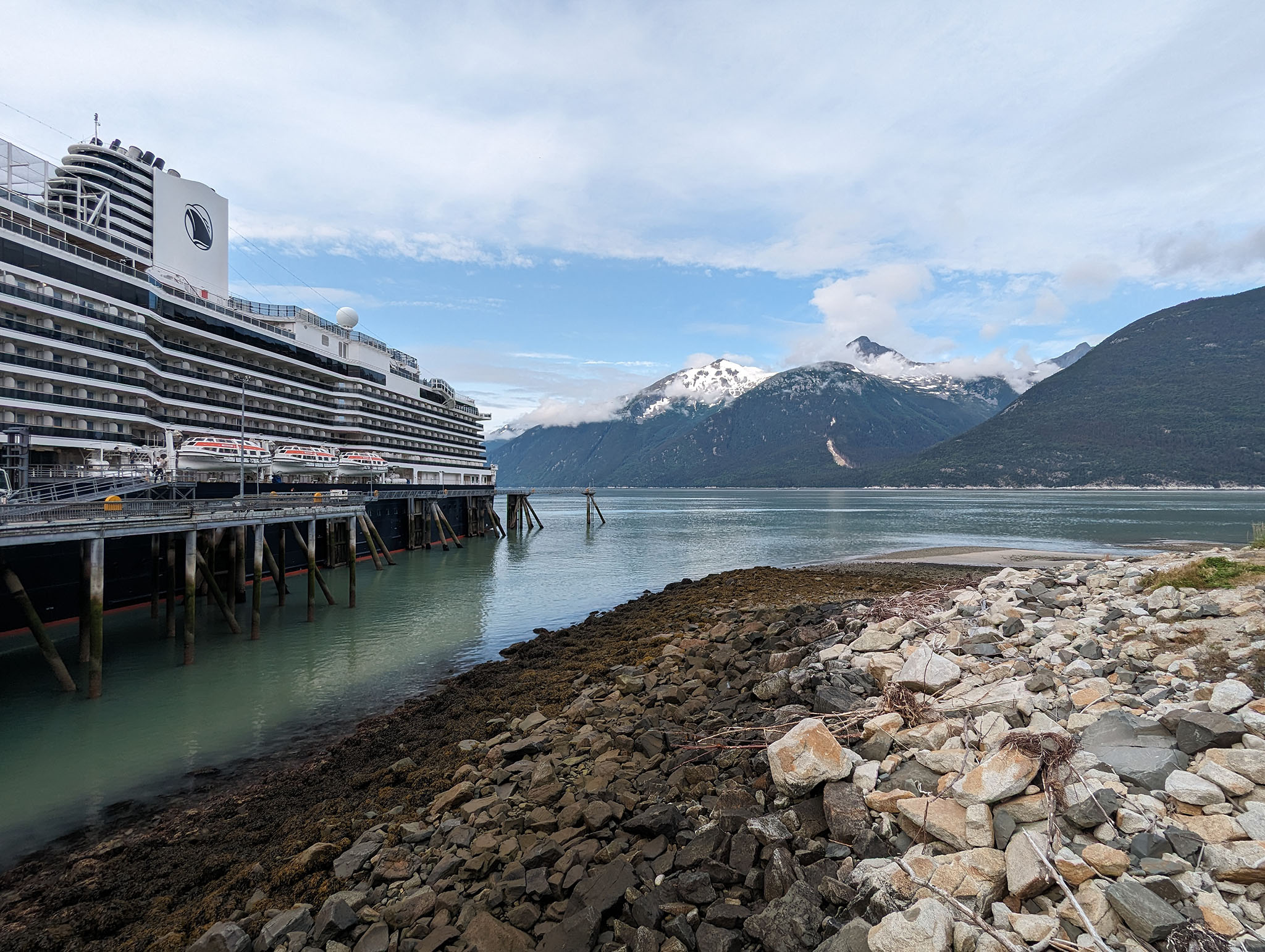 Rocky shore with flat water ahead of snow-peaked mountains and the back half of a cruise ship docked to the left of view