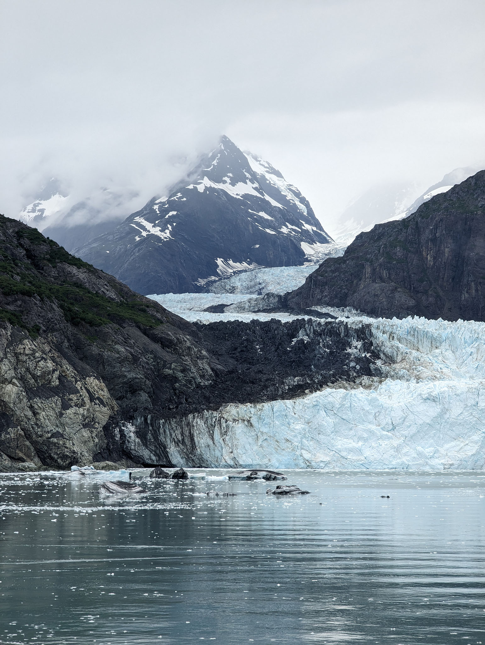 Icy cold water leading up to dark rocky cliffs and a blue-white glacier wall snaking up between mountains into the distance where a tall, snow-topped mountain is partly covered in low cloud