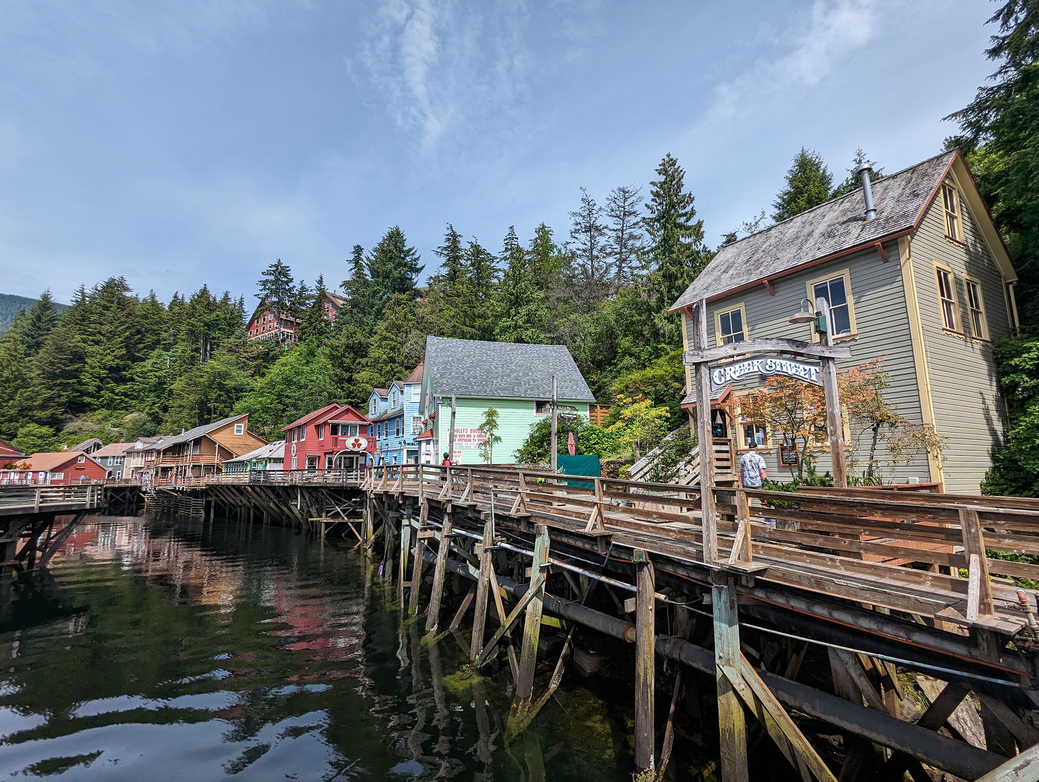 A wooden street raised on stilts over water with brightly coloured old buildings along the edge and a line of tall trees behind